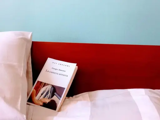Un libro aperto, "La camera azzurra" di Simenon, giace su un letto bianco, pronto per essere letto.