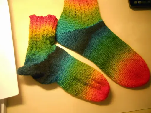 Två regnbågsfärgade, handstickade sockor i ett stycke. En större och en mindre, med färger som går från rosa och gult till grönt och blått.