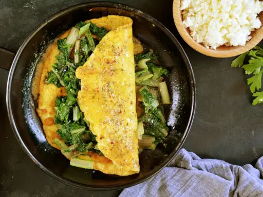Omlet z serka wiejskiego z zielonymi warzywami na patelni. Obok miseczka z serkiem i natka pietruszki.