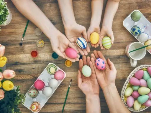 Hände von Kindern und Erwachsenen bemalen bunte Ostereier. Ein Korb voller Eier und Tulpen auf Holztisch.
