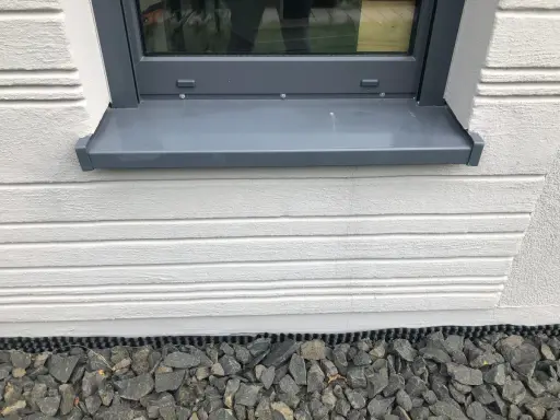 Graue Fensterbank mit leichter Neigung, die Regenwasser ableitet. Darunter weiße Holzverkleidung und grauer Schotter.