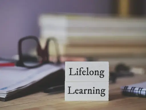 Dwa białe klocki z napisami "Lifelong" i "Learning" symbolizują uczenie się przez całe życie. W tle okulary i notatnik.