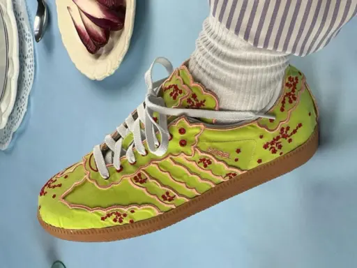 Jaskrawozielone buty Adidas z haftowanymi kwiatami wiśni i różowymi lamówkami, prawdopodobnie najdroższe buty Adidas, prezentowane na błękitnym tle.