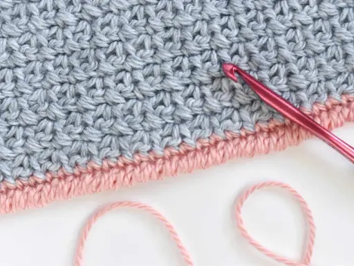 Detalle de tejido a crochet con punto cangrejo en rosa y gris. Una aguja rosa está lista para continuar el trabajo.