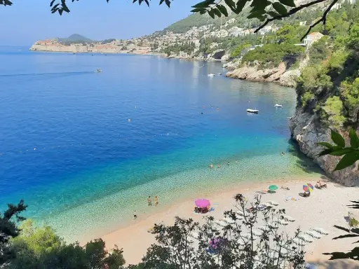 Malownicza zatoka z turkusową wodą i kamienistą plażą. W oddali widać mury Starego Miasta Dubrownik. Idealne plaże Dubrownik na relaks.