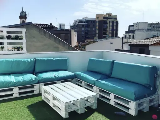 Sofá y mesa de centro hechos con palets blancos, con cojines turquesas, en una terraza urbana. ¡Una idea genial de como hacer un sofa con palets!