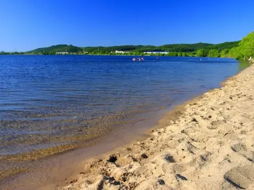Słoneczny dzień nad zalewem Pilica. Piaszczysta plaża, spokojna woda i zielone wzgórza w tle. Kilka osób pływa i odpoczywa.
