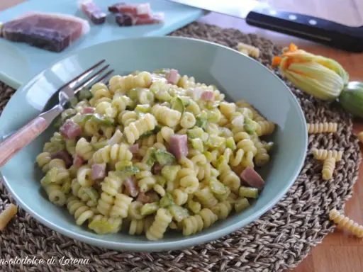 Piatto di pasta fusilli con zucchine e cubetti di speck, guarnito con un fiore di zucca e ingredienti freschi sul tagliere.