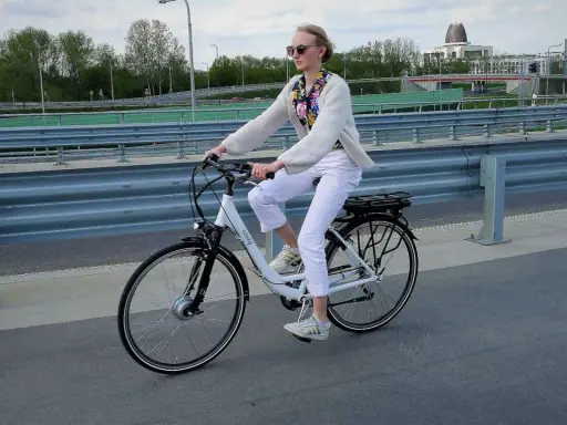 Młoda kobieta w białych spodniach i jasnym swetrze jedzie na rowerze elektrycznym, ciesząc się jazdą i osiągając swoją maksymalną prędkość.