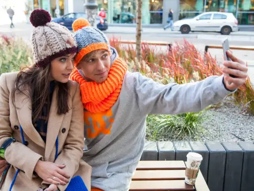 Para do selfie w zimowych stylizacjach. Ona w beżowym płaszczu i czapce z pomponem, on w szarej bluzie i czapce z pomarańczowym akcentem. Jaka czapka do płaszcza? Ta jej wygląda świetnie!