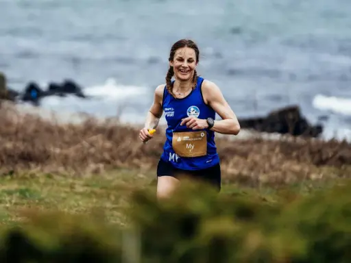 Woman running on a grassy trail by the sea, smiling.