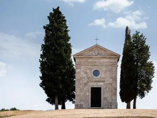 Piccola chiesa in pietra con cipressi, icona del Val d'Orcia, sotto un cielo azzurro con nuvole bianche.