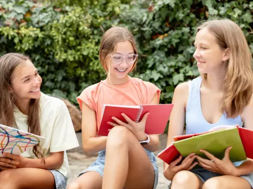 Tre ragazze sorridono studiando insieme, circondate dal verde. Usano quaderni colorati, ottime risorse didattiche Pearson per imparare all'aperto.