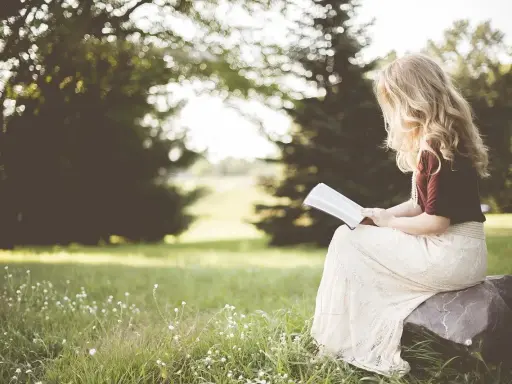Kobieta z blond włosami czyta psalm 6 tekst, siedząc na kamieniu w trawie, otoczona zielenią.