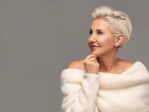 Le visage d'une femme de 80 ans, aux cheveux blonds courts, porte un pull blanc en maille. Elle regarde sur le côté, la main sous le menton.