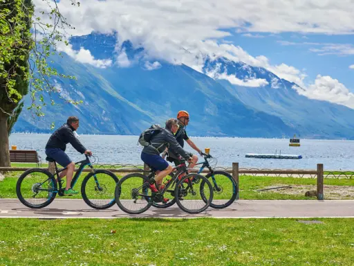 Ciclisti godono di una giornata soleggiata lungo uno dei laghi nord Italia, con montagne maestose sullo sfondo.