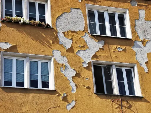Widoczne nierówności na elewacji budynku, gdzie żółty tynk odpada, odsłaniając białą warstwę. Cztery okna z białymi ramami.