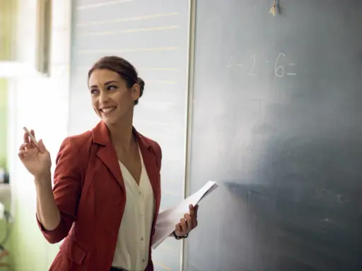 Lehrerin mit Kreide und Unterlagen vor einer Tafel mit einer Matheaufgabe. Sie lächelt und wirkt engagiert, was auf einen interaktiven Unterricht hindeutet.