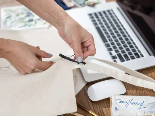 Ręce tworzą torbę DIY, przyszywając czarną wstążkę do płóciennego materiału. Obok leży laptop i myszka.