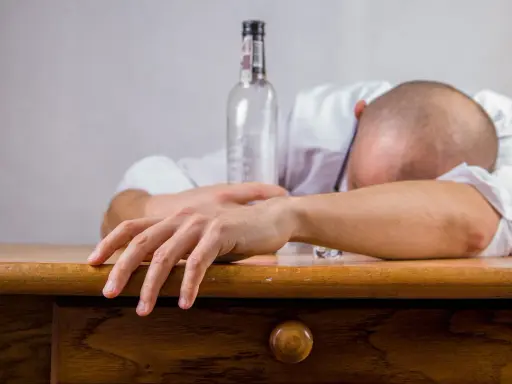 Mężczyzna w białej koszuli leży na stole, głowa oparta na ramieniu. Obok pusta butelka, symbolizująca skutki spożycia alkoholu, jakimi mogą być wymioty po alkoholu.