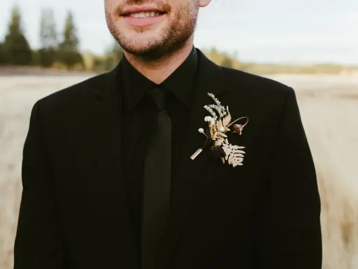 A groom in all black wedding guest attire men's suit, complete with a black tie and a rustic boutonniere.