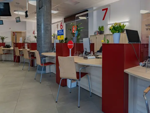 Puste biurka z krzesłami w urzędzie, gdzie przestrzegana jest proksemika. Widać numery stanowisk i tabliczki informacyjne.