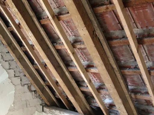 Charpente en bois avec des tuiles rouges et un mur en briques décrépit. Les contre latte sont visibles, soutenant la structure.