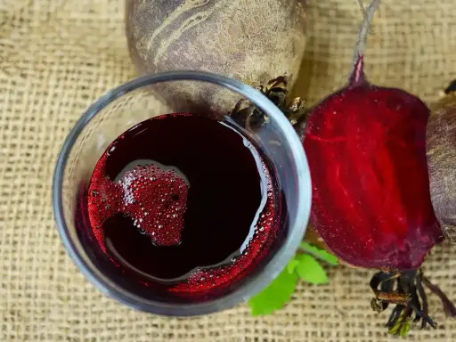 Verre de jus de betterave rouge vif, avec des betteraves entières et coupées autour. Une boisson saine pour savoir comment faire du jus de betterave.
