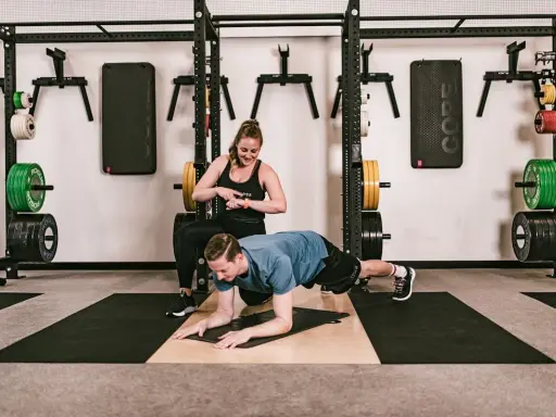Ein Mann macht einen Unterarmstütz, während eine Trainerin die Zeit stoppt. Perfekt für einen HIIT Trainingsplan im Fitnessstudio.