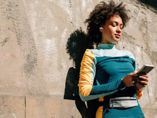 Frau mit Kopfhörern lehnt an Wand, hält handy beim joggen. Musik und Motivation für die Laufstrecke.