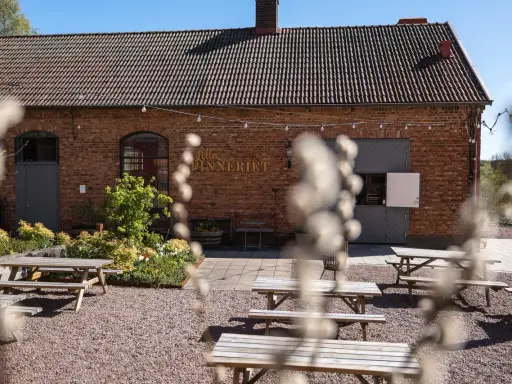 Utomhusmiljö vid Lilla Spinneriet med tegelbyggnad, trädgård och flera picknickbord.