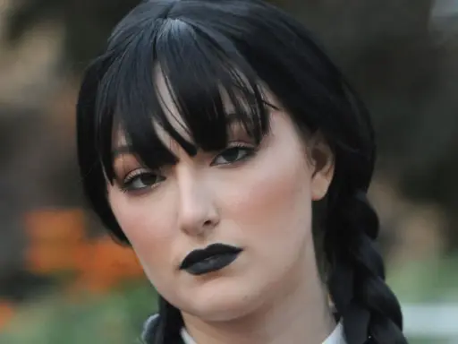 Maquillaje de Miércoles Addams con labios negros y ojos ahumados, peinado con trenzas y flequillo.