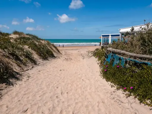 Sandiger Weg zum Conil de la Frontera Strand, mit Dünen, Meer und einem Strandcafé im Hintergrund.