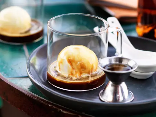 Ein cremiger Affogato mit Alkohol, serviert in einem Glas mit einer Kugel Vanilleeis, übergossen mit Espresso und einem kleinen Schnapsglas daneben.