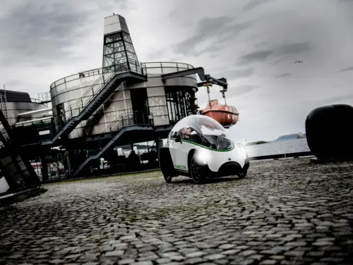 Ein Podbike Frikar fährt auf Kopfsteinpflaster vor einem modernen Gebäude am Wasser.