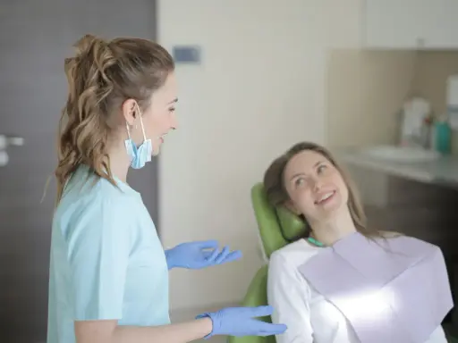 Dentystka w niebieskiej bluzie i rękawiczkach rozmawia z pacjentką. Długie studia stomatologiczne wymagają zaangażowania.