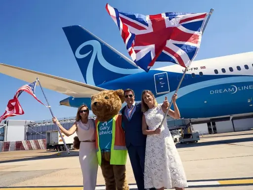Två kvinnor och en man med en björndräkt firar framför ett flygplan från Norse Atlantic Airways. En kvinna håller en amerikansk flagga, en annan en brittisk flagga.