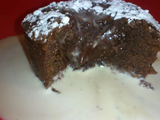 Un fondant au chocolat dégoulinant, saupoudré de sucre glace, servi dans une sauce crémeuse sur une assiette rouge.