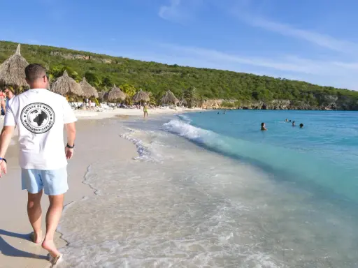 En man går längs en vit sandstrand med turkost vatten. Palmklippor och parasoll syns i bakgrunden. En perfekt plats att undra var ligger Curacao.