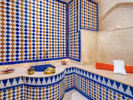 Ein luxuriöser Hamam in Marokko, verziert mit blauen und orangen Zellige-Fliesen. Kerzen und Schalen mit Rosenblättern schaffen eine entspannende Atmosphäre.