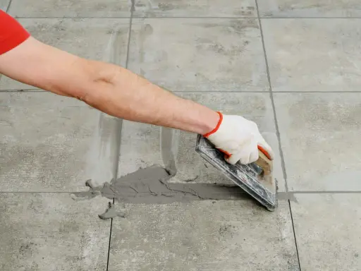 Mano con guante aplica lechada gris a las juntas entre azulejos de cemento con una llana.