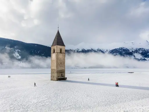 Torre sommersa nel lago di Resia, circondata da neve e montagne nebbiose. Alcune persone passeggiano sul ghiaccio.