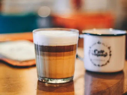 Ein geschichteter Latte Macchiato im Glas, daneben eine weiße Tasse mit Kaffee-Aufdruck.