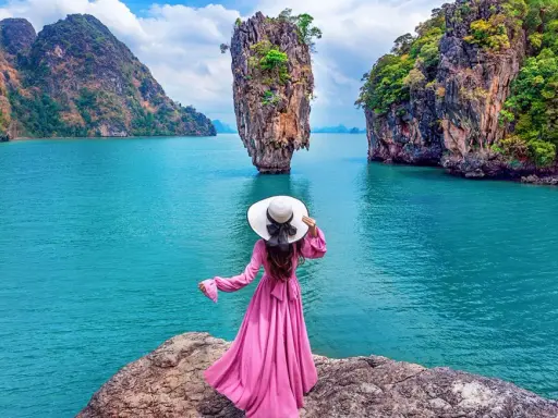 Donna con abito rosa e cappello bianco ammira la spettacolare **isola di James Bond** con le sue formazioni rocciose uniche e il mare turchese.