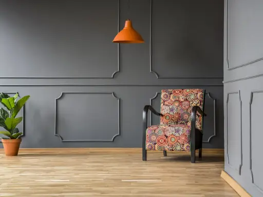 Nowoczesne sztukateria ścienna inspiracje: fotel w kolorowe wzory, pomarańczowa lampa i roślina na tle szarej ściany.