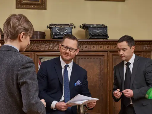 Czterech mężczyzn w garniturach rozmawia w gabinecie z zabytkowymi maszynami do pisania.