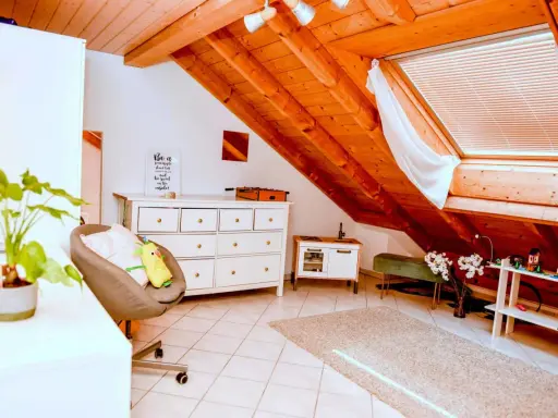 Acogedor despacho en buhardilla con vigas de madera, cómoda blanca, sillón y alfombra.