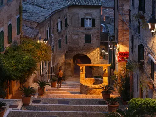 Antica scala a Gabicce Monte illuminata da lampioni, con un pozzo in mattoni e piante in vaso.