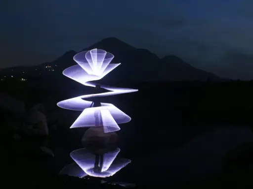 Un artista crea forme luminose con la light painting fotografia, disegnando spirali e petali di luce viola su uno sfondo scuro con montagne e riflessi sull'acqua.