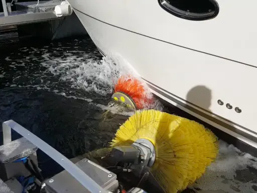Nettoyage de la partie immergée d'un bateau avec des brosses rotatives jaunes et oranges. L'eau éclabousse autour des brosses.